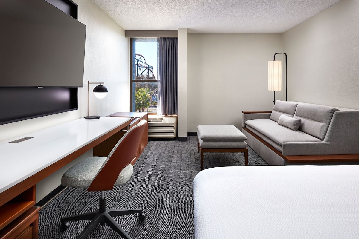 A hotel room with a large flat-screen TV, a desk with a chair, a sofa, an ottoman, and a lamp.