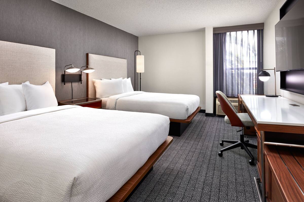 A hotel room with two beds, a desk, a chair, a TV, and two wall-mounted lamps.