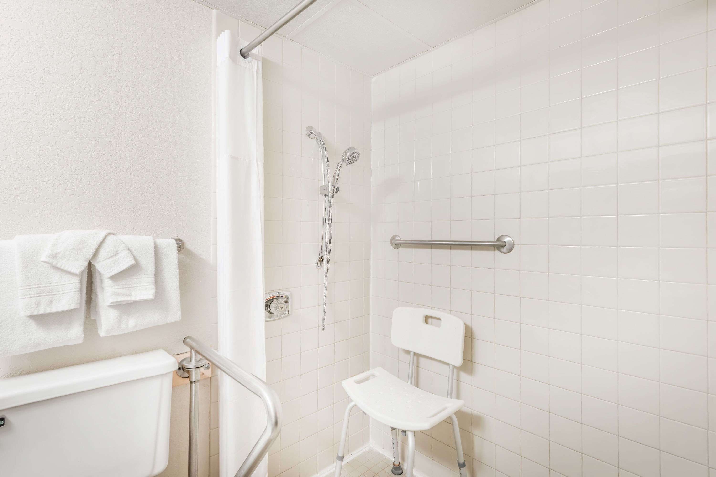 A bathroom with a white tiled wall, a shower with a handheld showerhead, a grab bar, a white toilet, and a white chair.