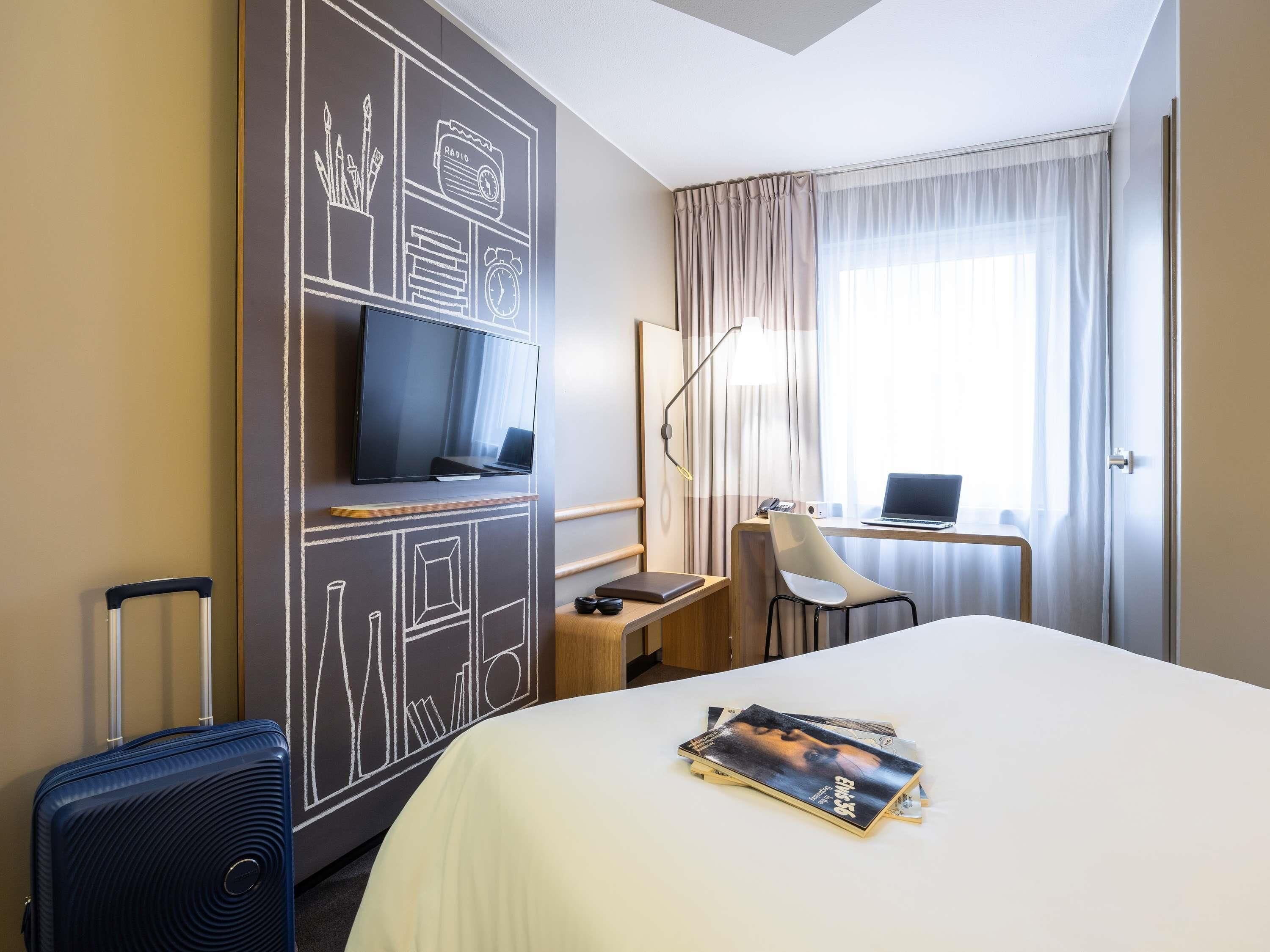 A hotel room with a bed, a desk with a laptop, a chair, a TV mounted on a wall, and a suitcase.