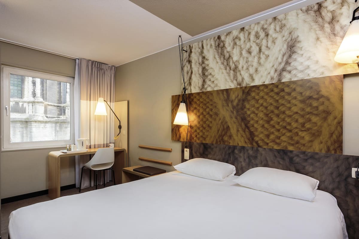 A hotel room with a bed, desk, chair, and a view of a building through a window.