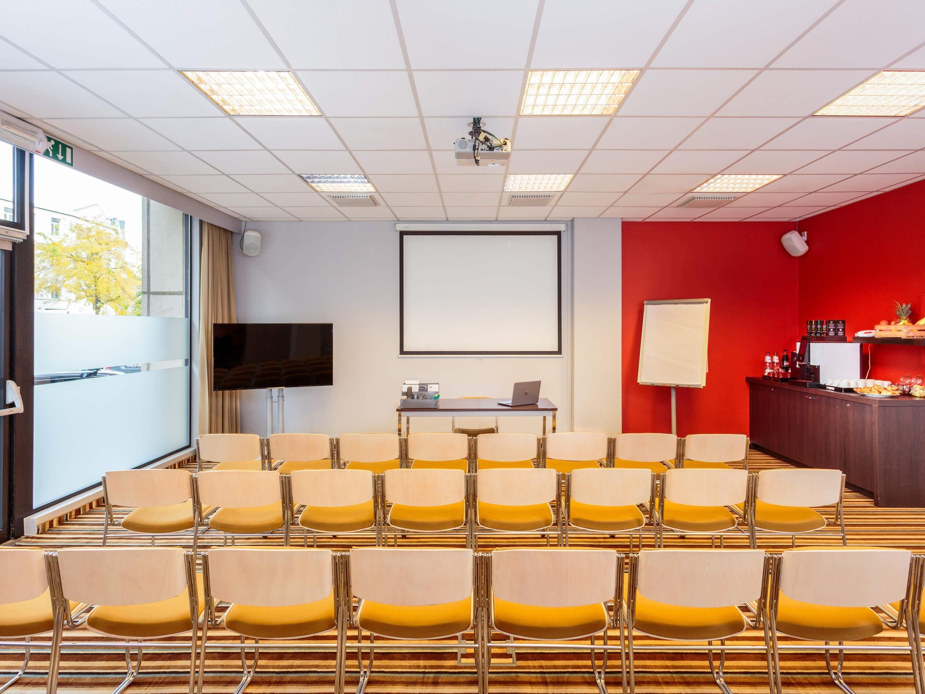 A conference room with rows of chairs, a projector screen, a whiteboard, and a desk with a laptop.