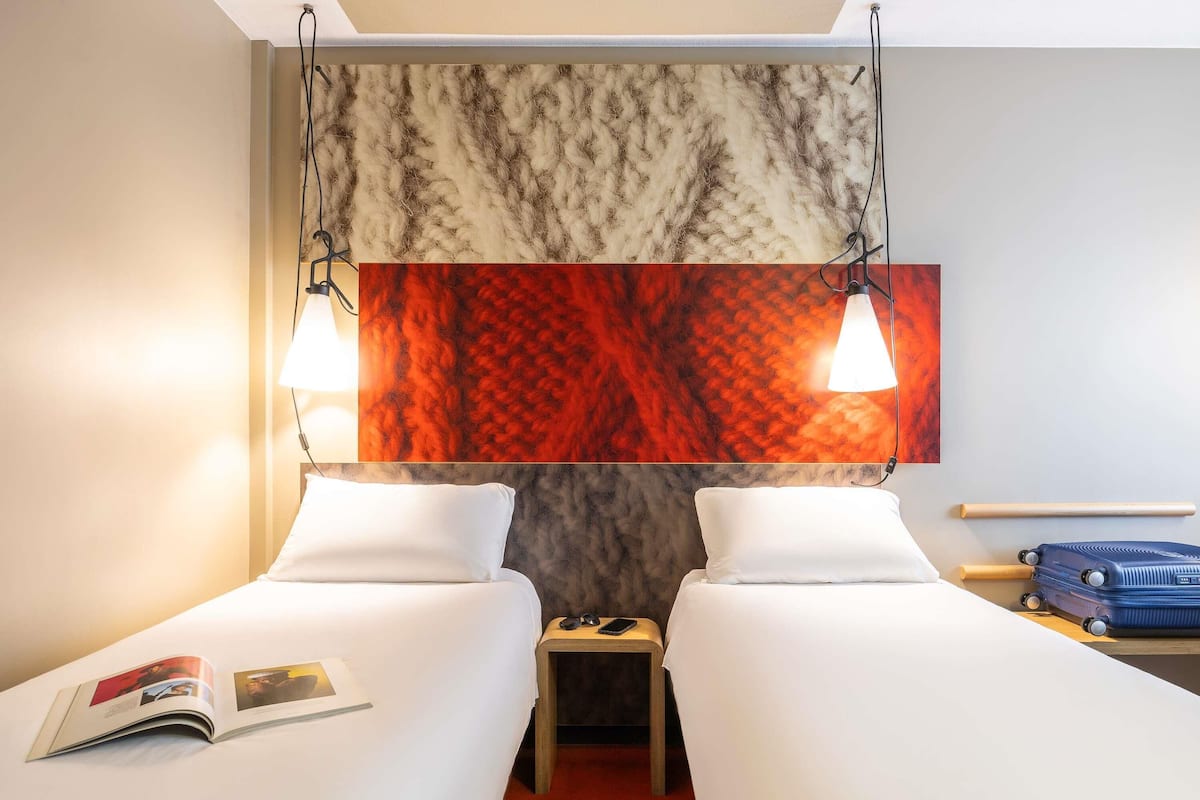 A hotel room with two beds, a red carpet, a small table with a lamp, a blue suitcase, and a magazine on the bed.
