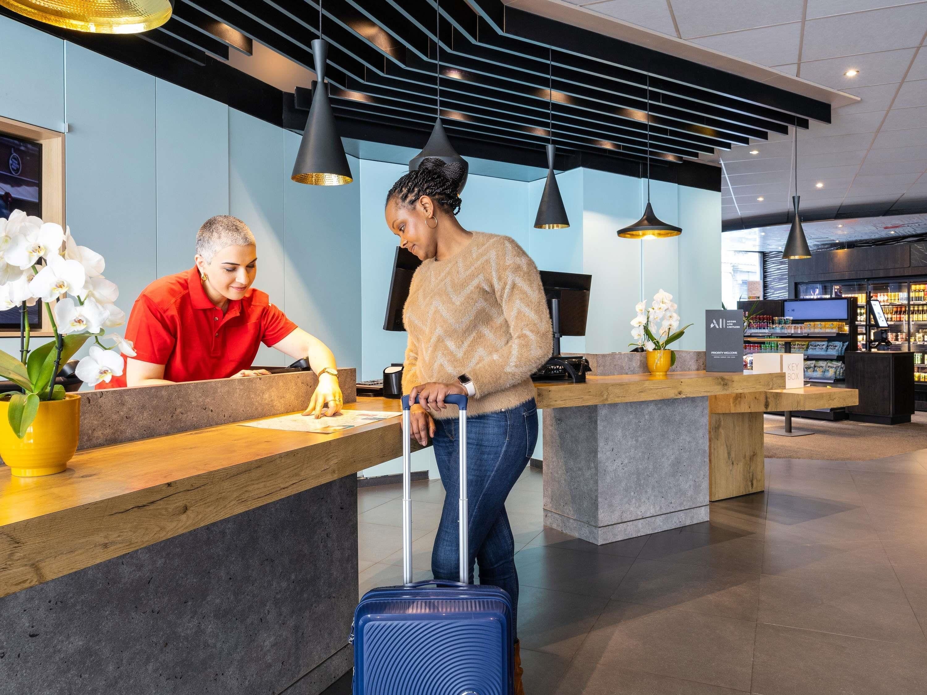 A reception desk with a counter, a person in a red shirt behind it, and another person with a suitcase in front.