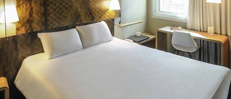 A hotel room with a large bed, a desk, and a chair. There is a window with curtains and a lamp on the desk.