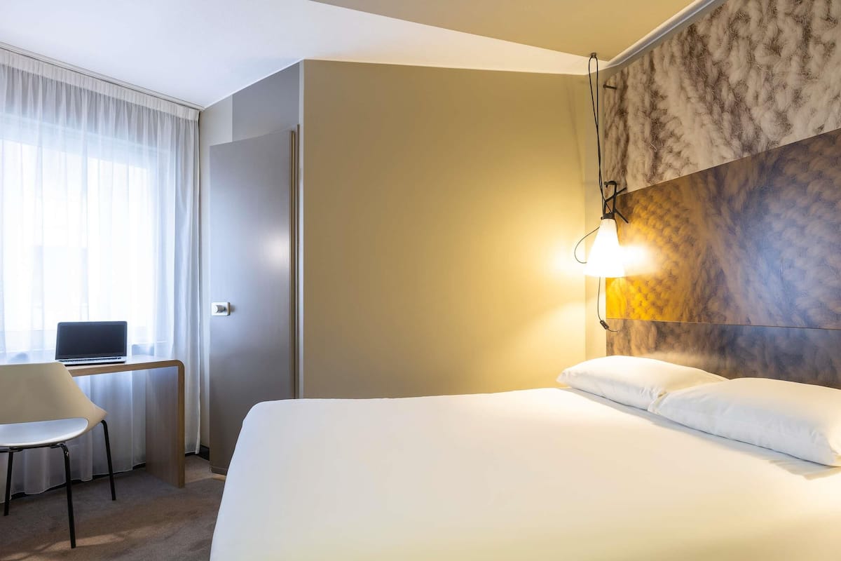 A hotel room with a bed, a desk with a laptop, a chair, and a wall-mounted lamp.