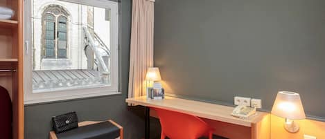 A hotel room with a desk, two chairs, a bench, a window with a view of a bridge, and a lamp.