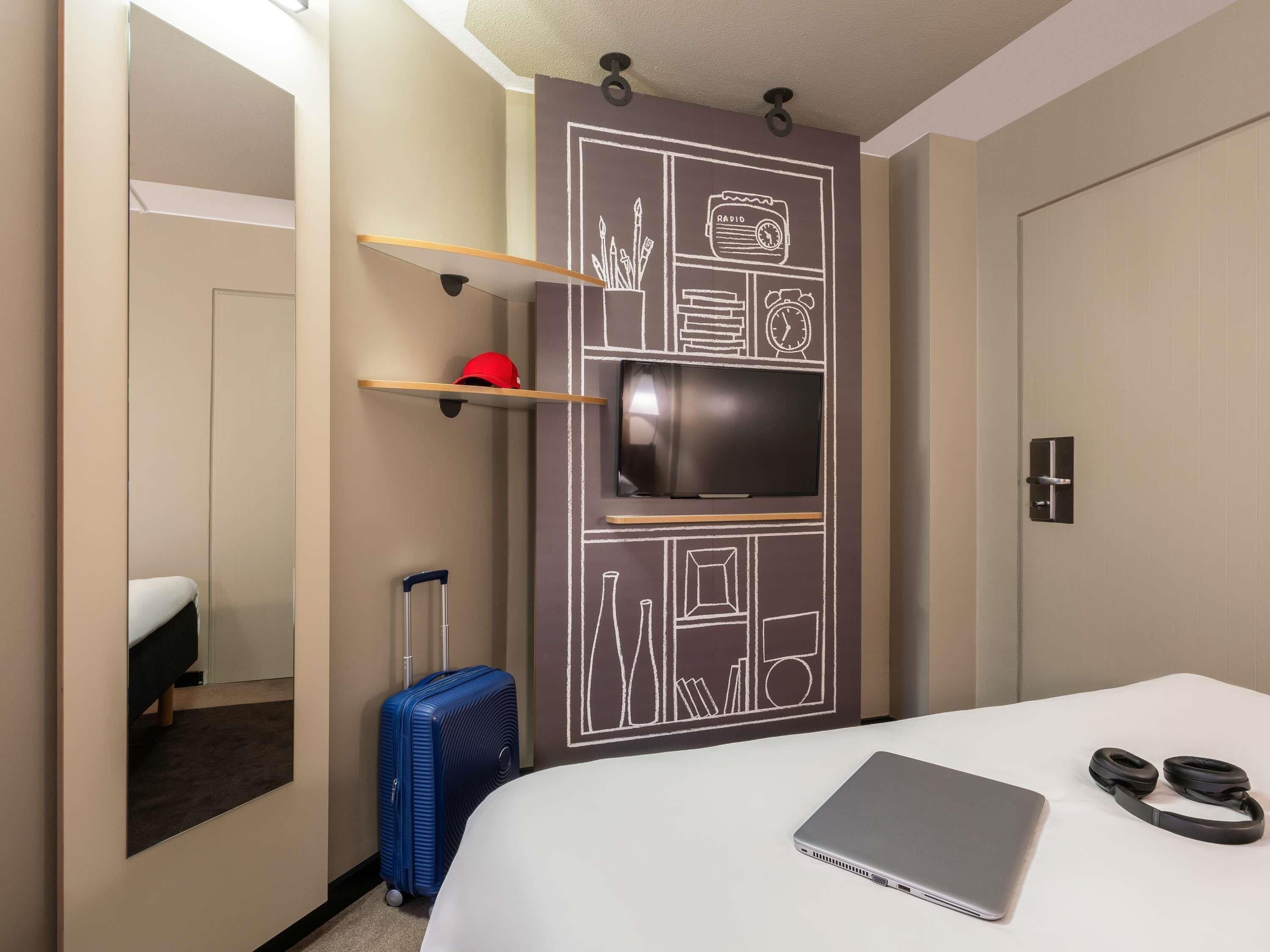 Une chambre d’hôtel équipée d’un lit, d’un ordinateur portable, d’écouteurs, d’une valise et d’un téléviseur fixé au mur, le tout dans un décor rappelant un tableau noir.
