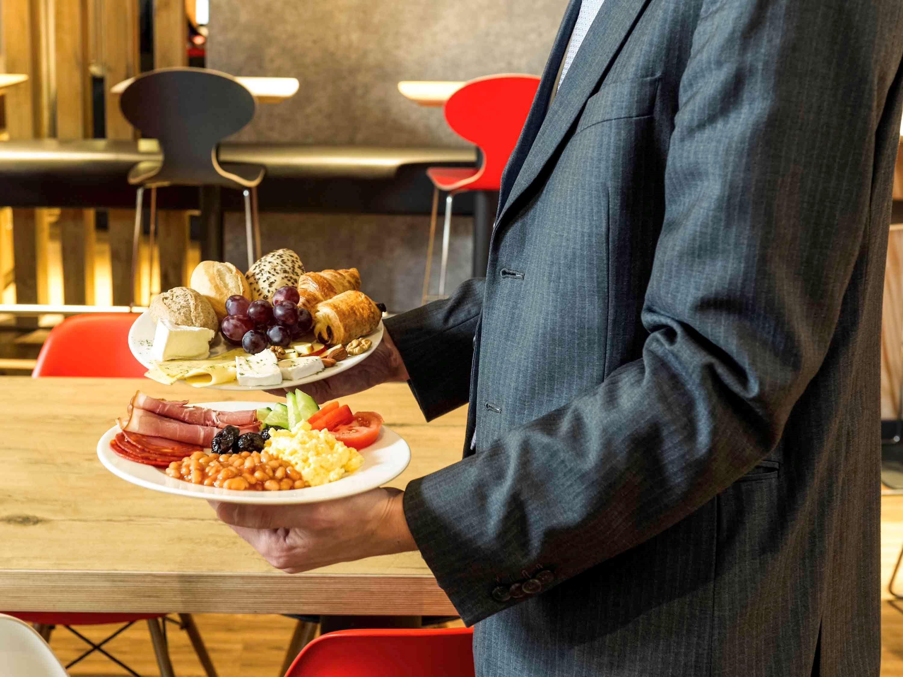 A person holding two plates of food in a modern dining area with wooden and red chairs.
