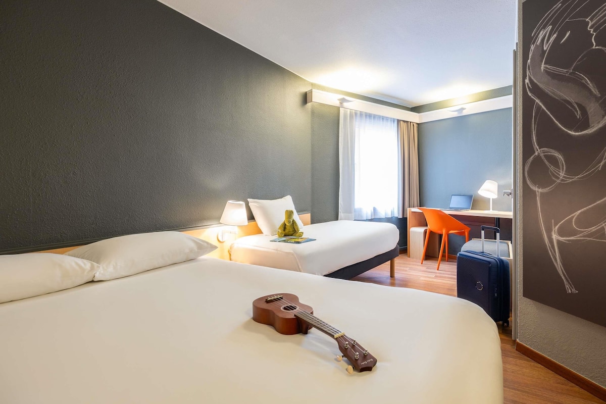 A hotel room with two beds, a desk, a chair, a guitar, and a wall with a chalk drawing.