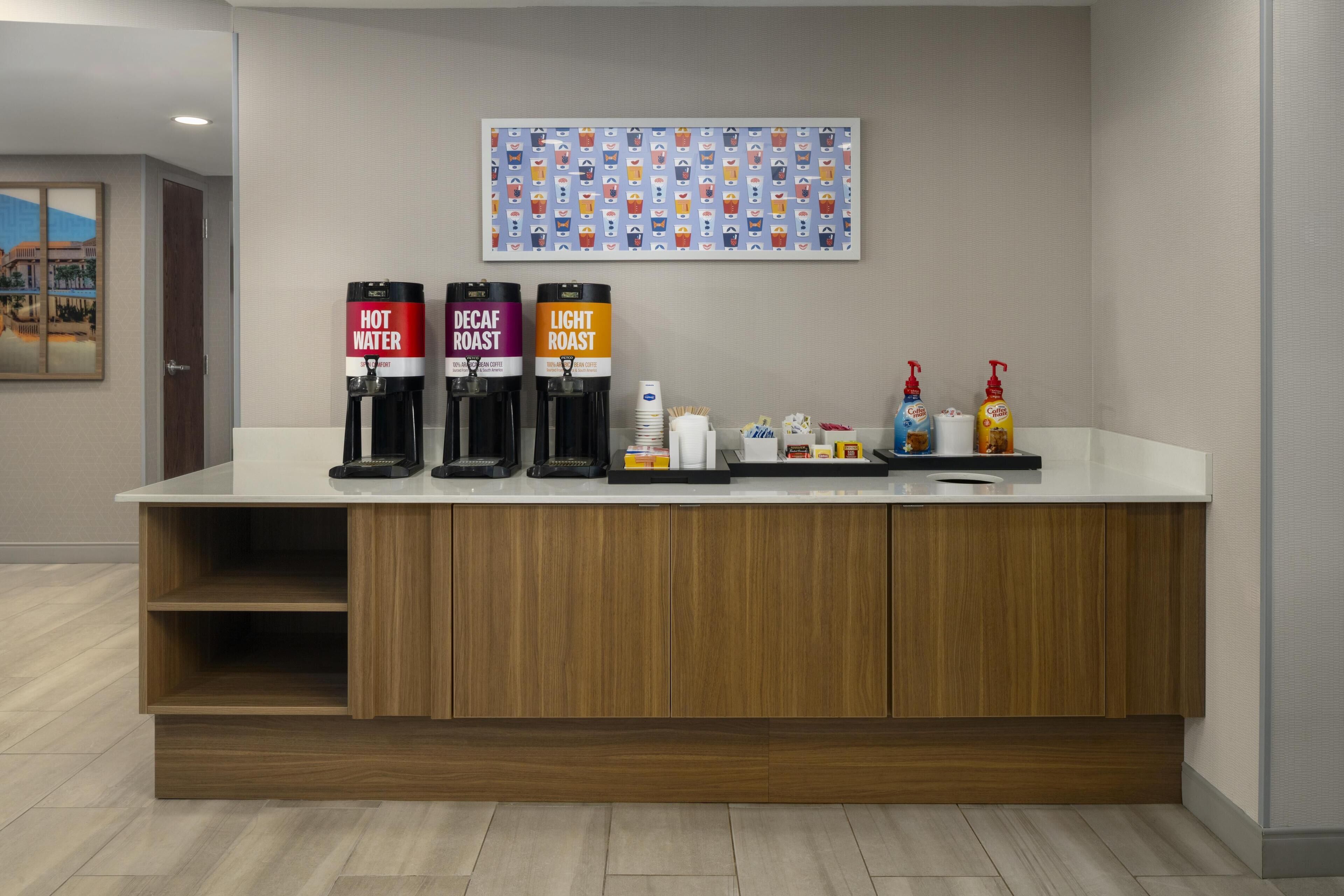 Zona de recepción de hotel con mostrador, máquinas de café de agua caliente, descafeinado y tueste claro, y artículos de aseo.