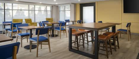 A modern dining area with multiple tables and chairs, large windows, and a flat-screen TV mounted on the wall.