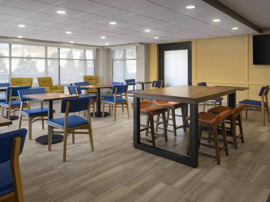 A modern dining area with multiple tables and chairs, large windows, and a flat-screen TV mounted on the wall.