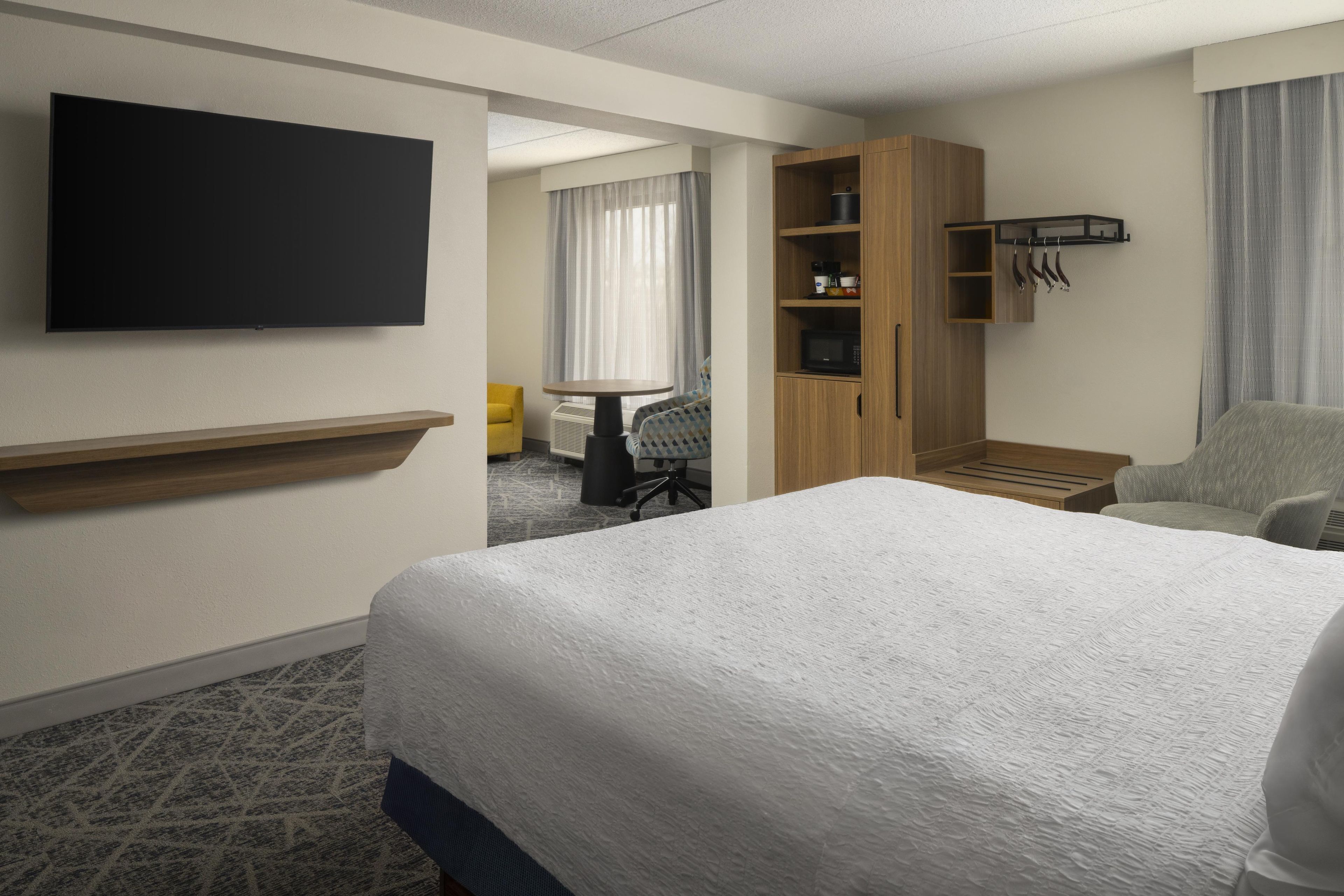 A hotel room with a large bed, a flat-screen TV mounted on the wall, a wooden wardrobe, a small table with a chair, and a seating area with a yellow chair and a window with curtains.