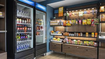 A vending machine stocked with beverages and snacks, with a wooden shelf unit displaying various packaged goods.