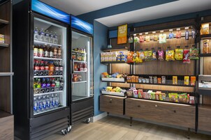 Máquina expendedora repleta de bebidas y aperitivos, con un estante de madera que exhibe diversos productos envasados.