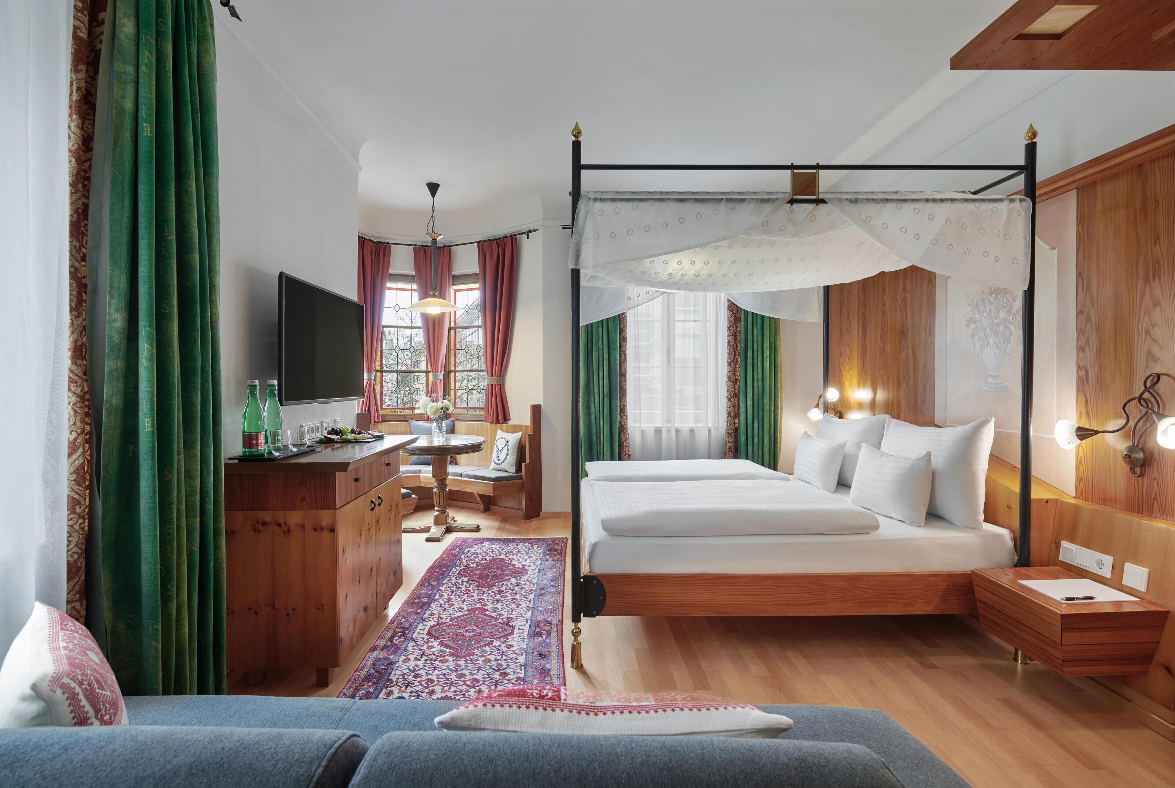 A bedroom with a four-poster bed, a wooden dresser, a television, a desk with a chair, and a window with curtains.