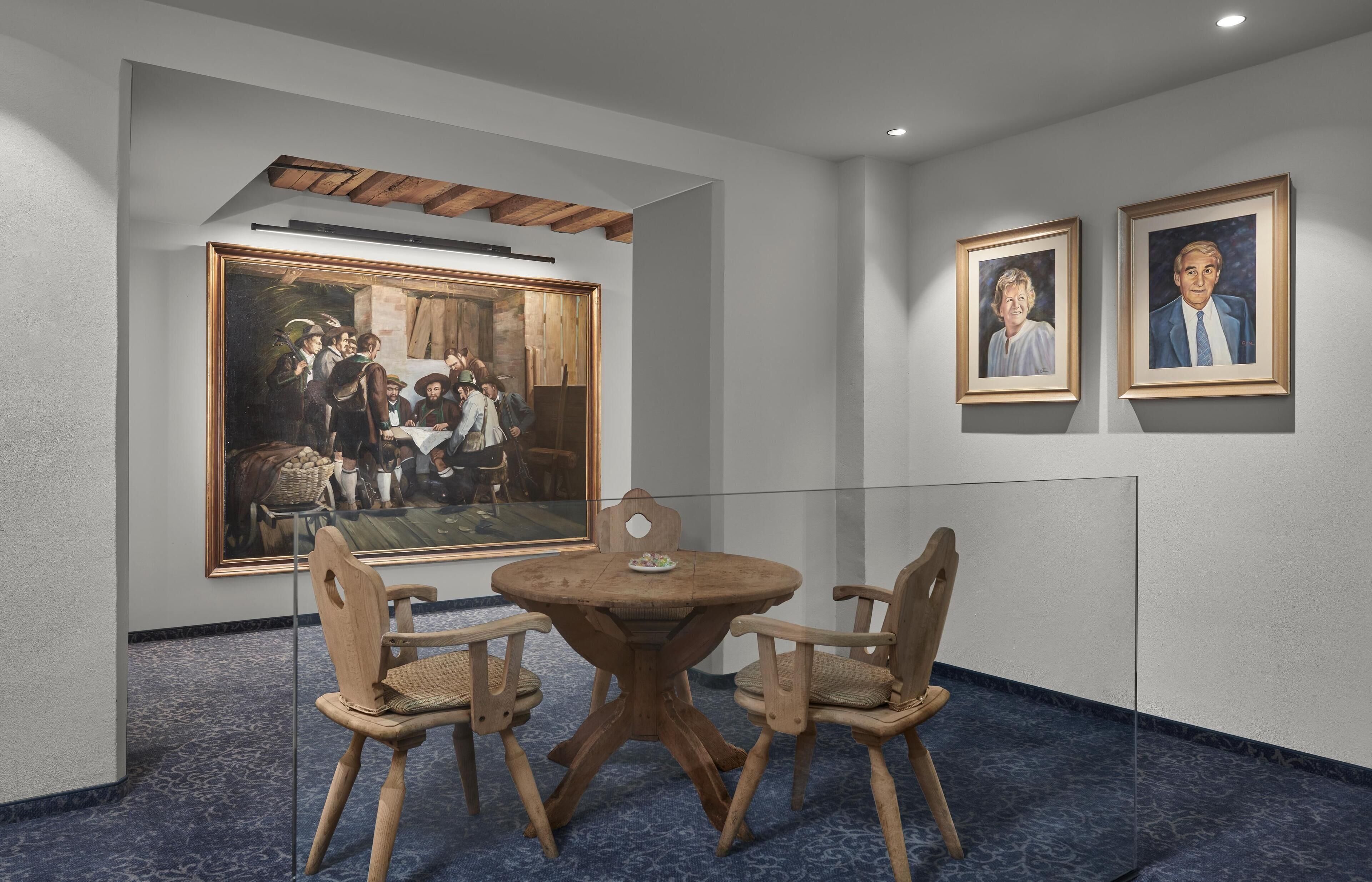 A dining area with a round wooden table and four chairs, a large framed painting on the wall, and two smaller framed portraits.