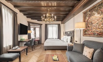 Una habitación de hotel con una cama grande, un sofá, un comedor con mesa y sillas, un televisor y una araña.