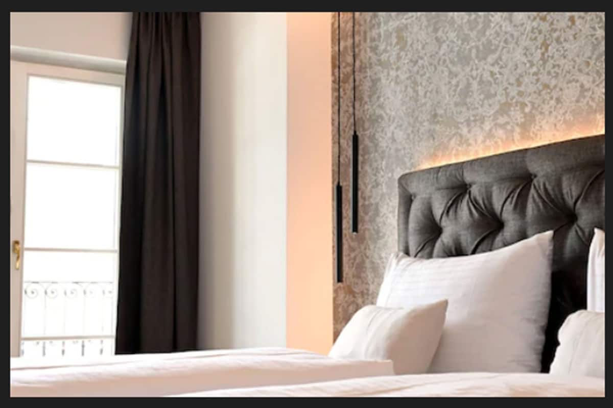 Une chambre à coucher avec une tête de lit capitonnée, une grande fenêtre avec des rideaux et un mur orné d’un motif.