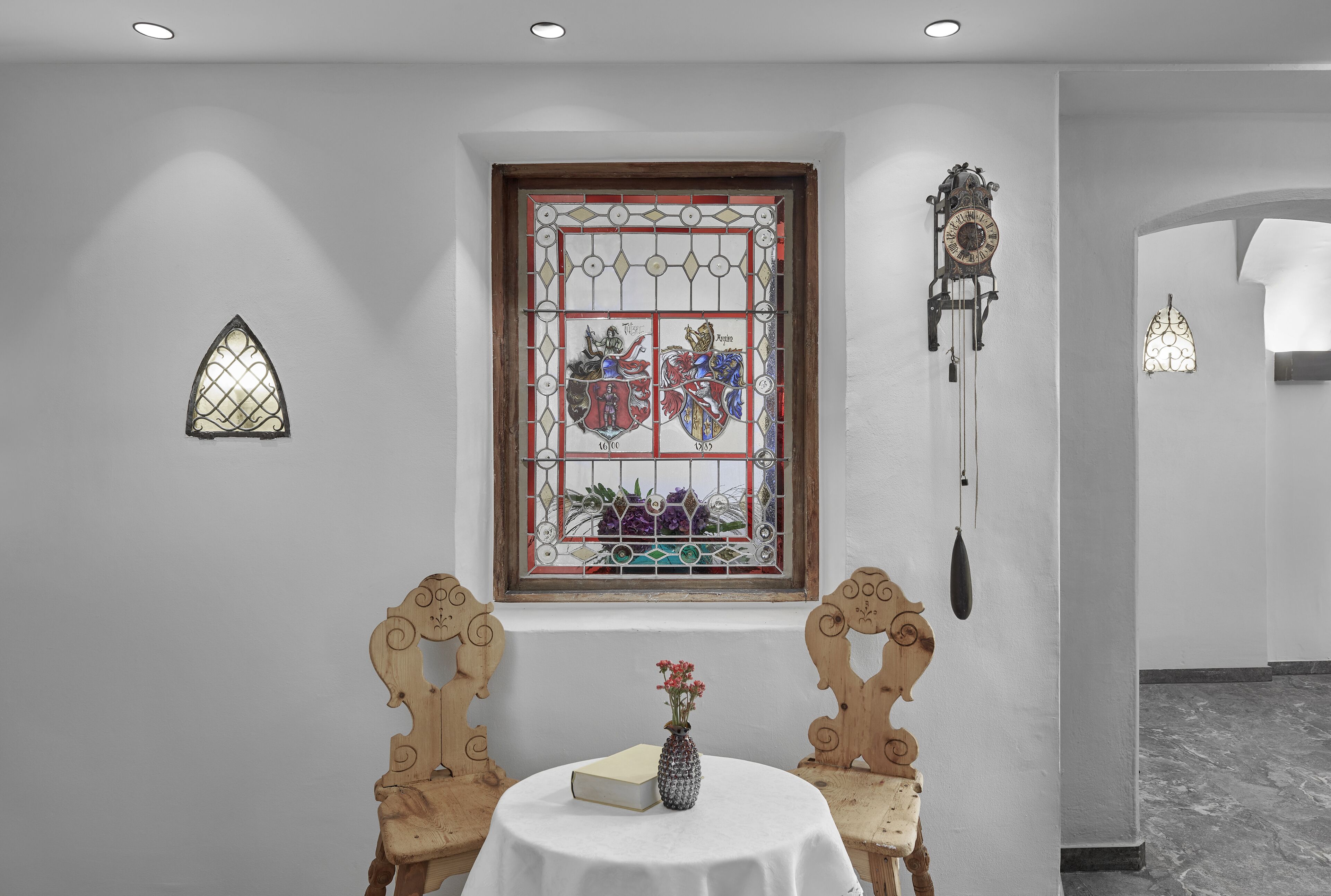 A room with a stained glass window, a wooden chair, a table with a book and vase, and a wall-mounted clock.