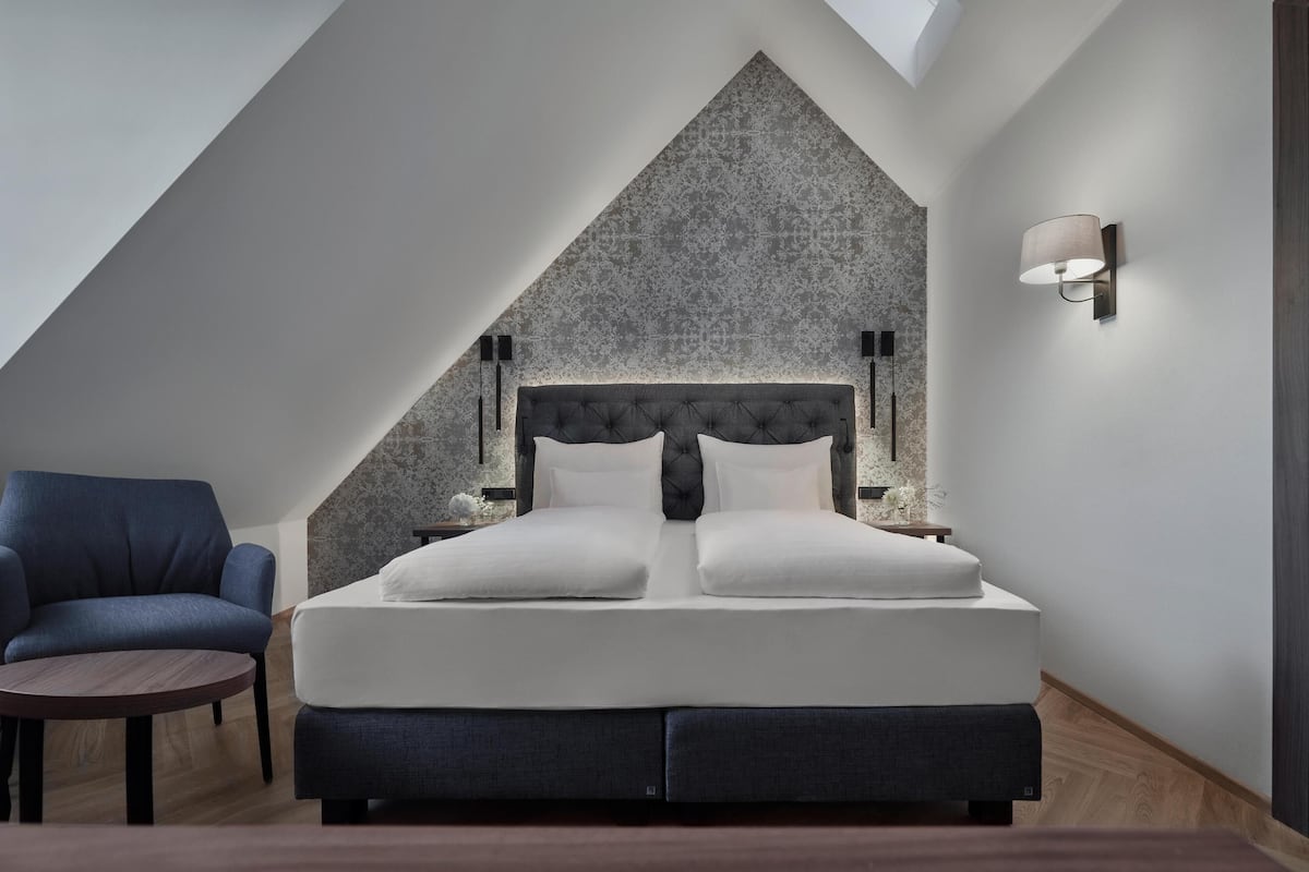 Une chambre à coucher moderne avec un plafond haut, un grand lit doté d’une tête de lit capitonnée, un fauteuil bleu, une table ronde en bois et un mur à texture.