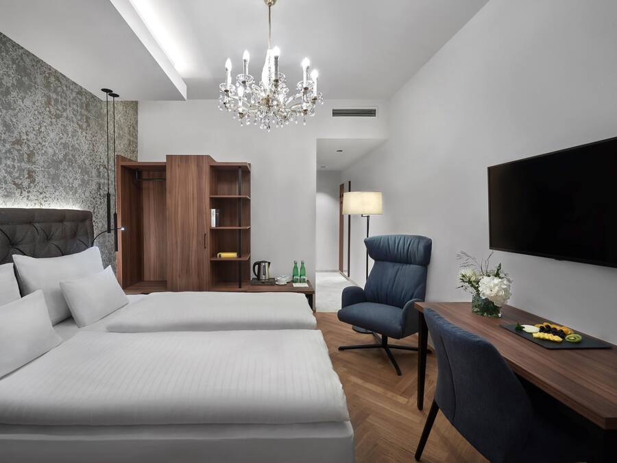 A modern hotel room with a large bed, a desk, a chair, a TV, a chandelier, and a bookshelf.