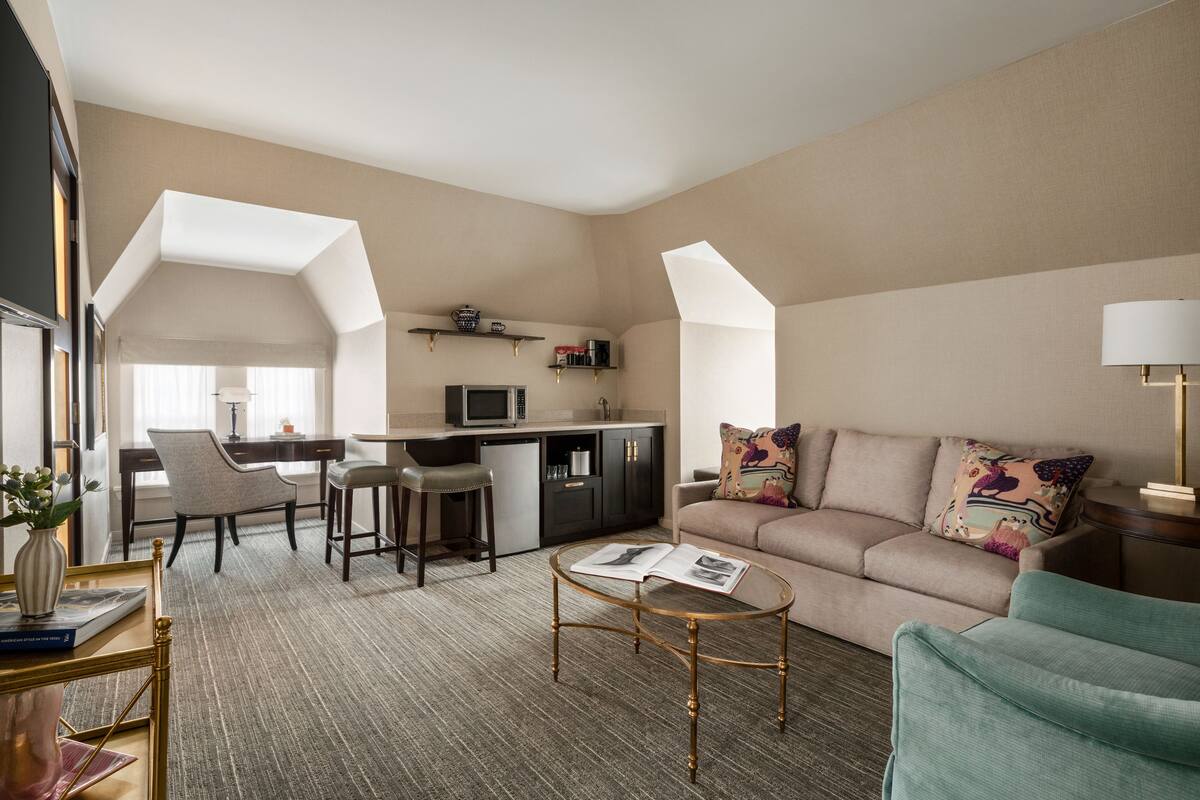 A modern hotel room with a kitchenette, a sofa, a dining area, and a large window providing natural light.
