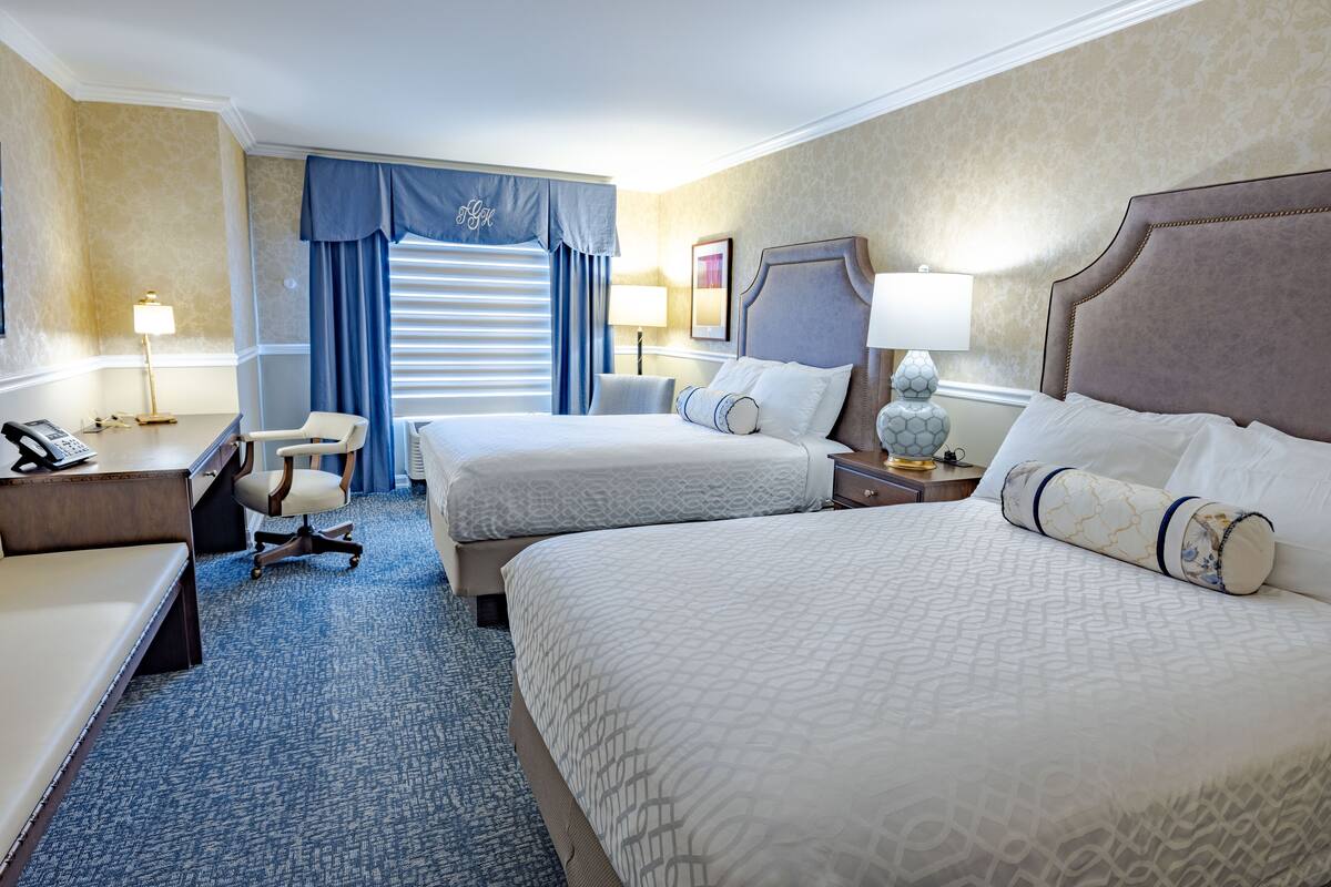 A hotel room with two beds, a desk, a chair, and a lamp.