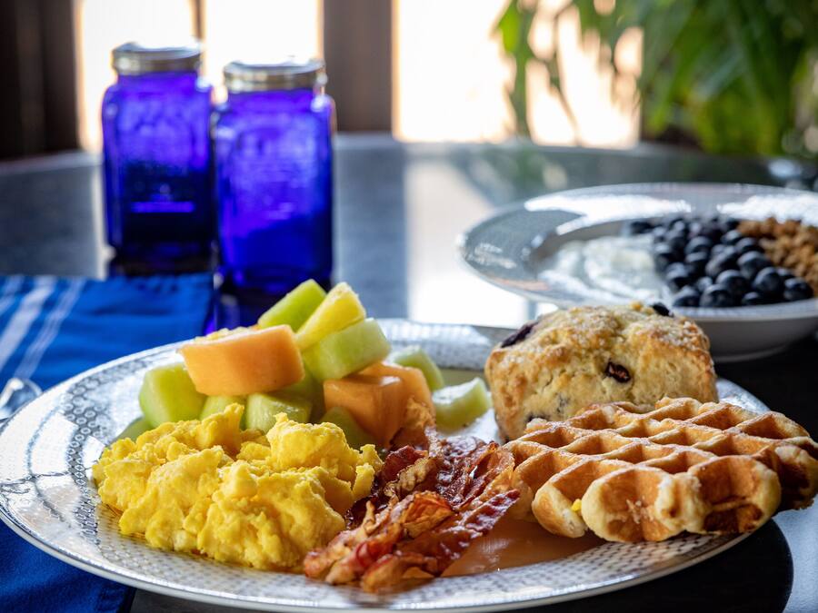 Une assiette de petit-déjeuner contenant des œufs brouillés, du bacon, des gaufres et des fruits, posée sur une table recouverte d’une nappe bleue.