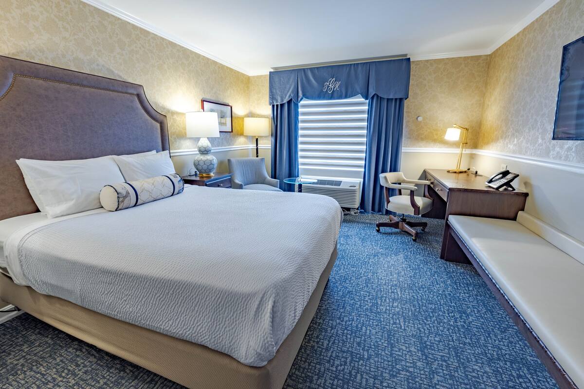 A hotel room with a large bed, a desk, a chair, a lamp, and a window with curtains.