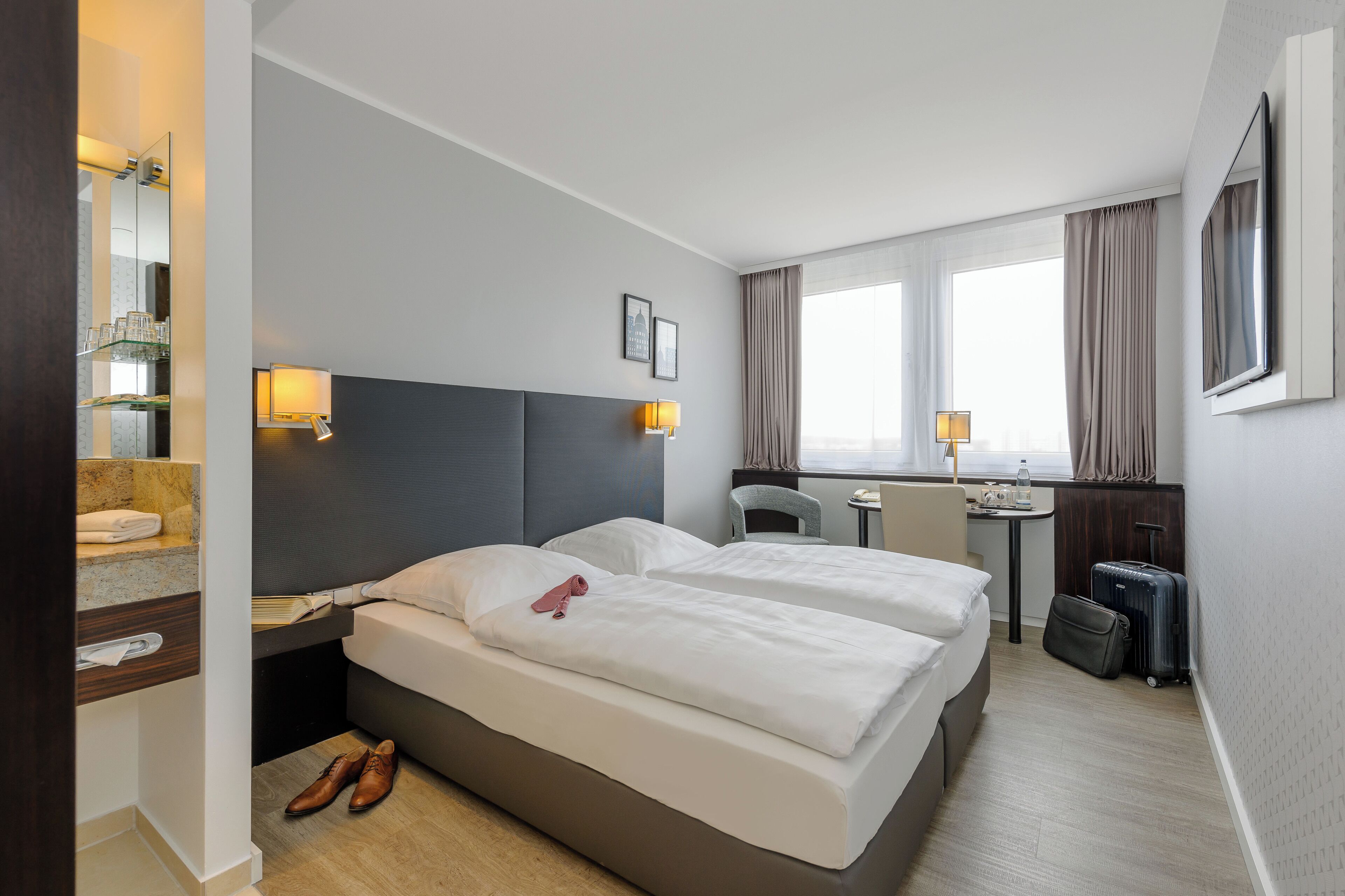 Habitación de hotel con dos camas, un escritorio, una silla, una mesita, un espejo y una ventana con cortinas.