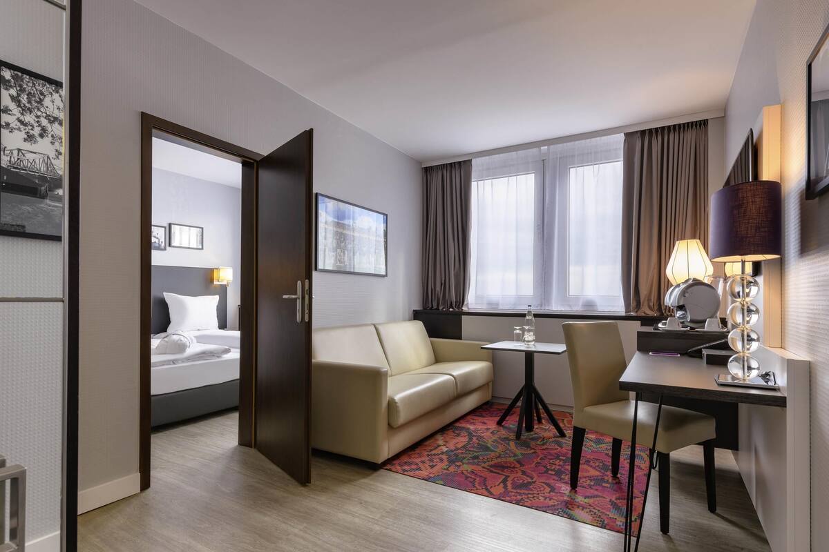 A hotel room with a bed, a sofa, a desk, a chair, a small table, and a window with curtains.