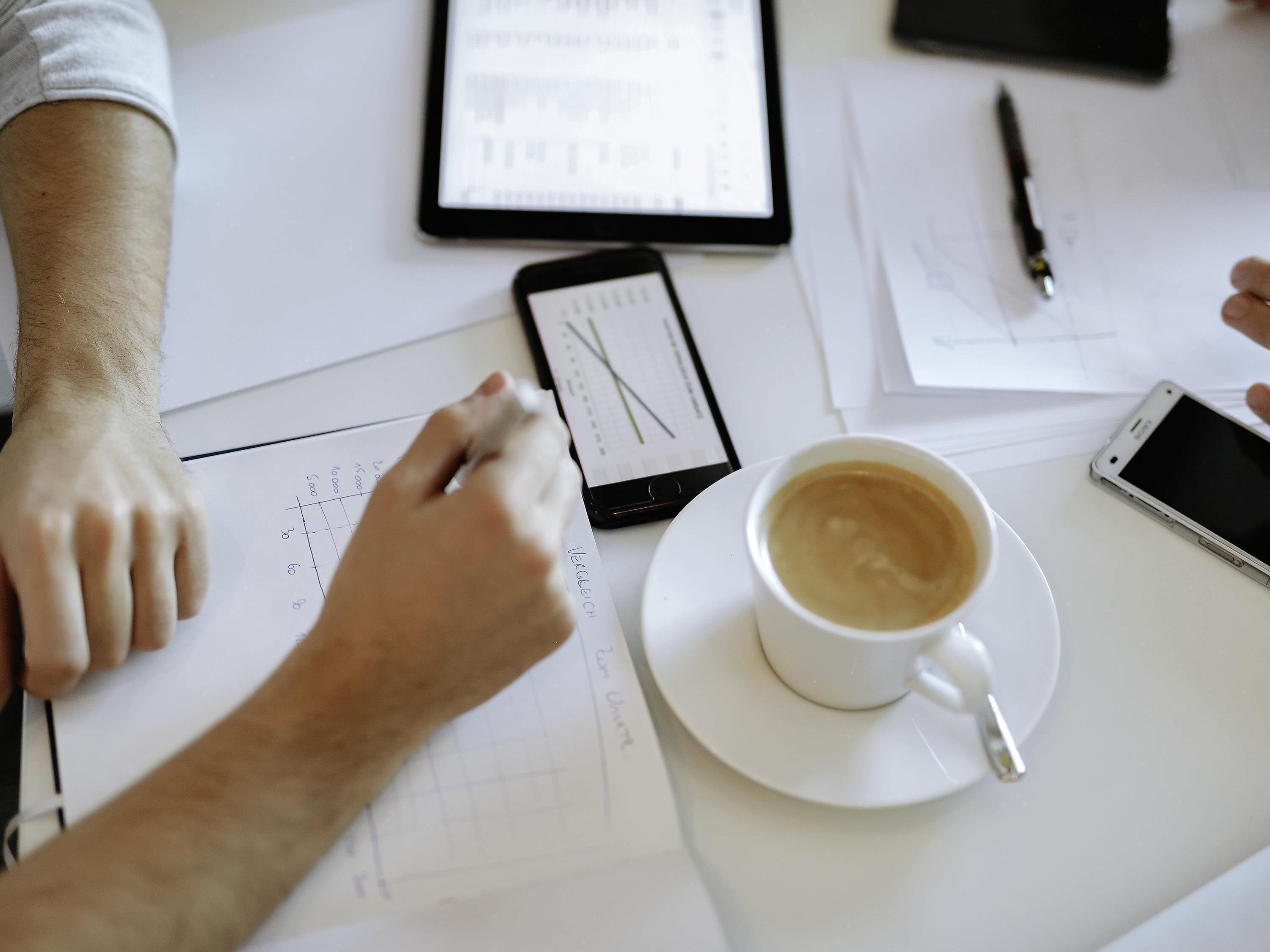 En person som sitter vid ett skrivbord med papper, en smartphone, en penna och en kopp kaffe.