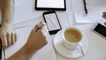 En person som sitter vid ett skrivbord med papper, en smartphone, en penna och en kopp kaffe.