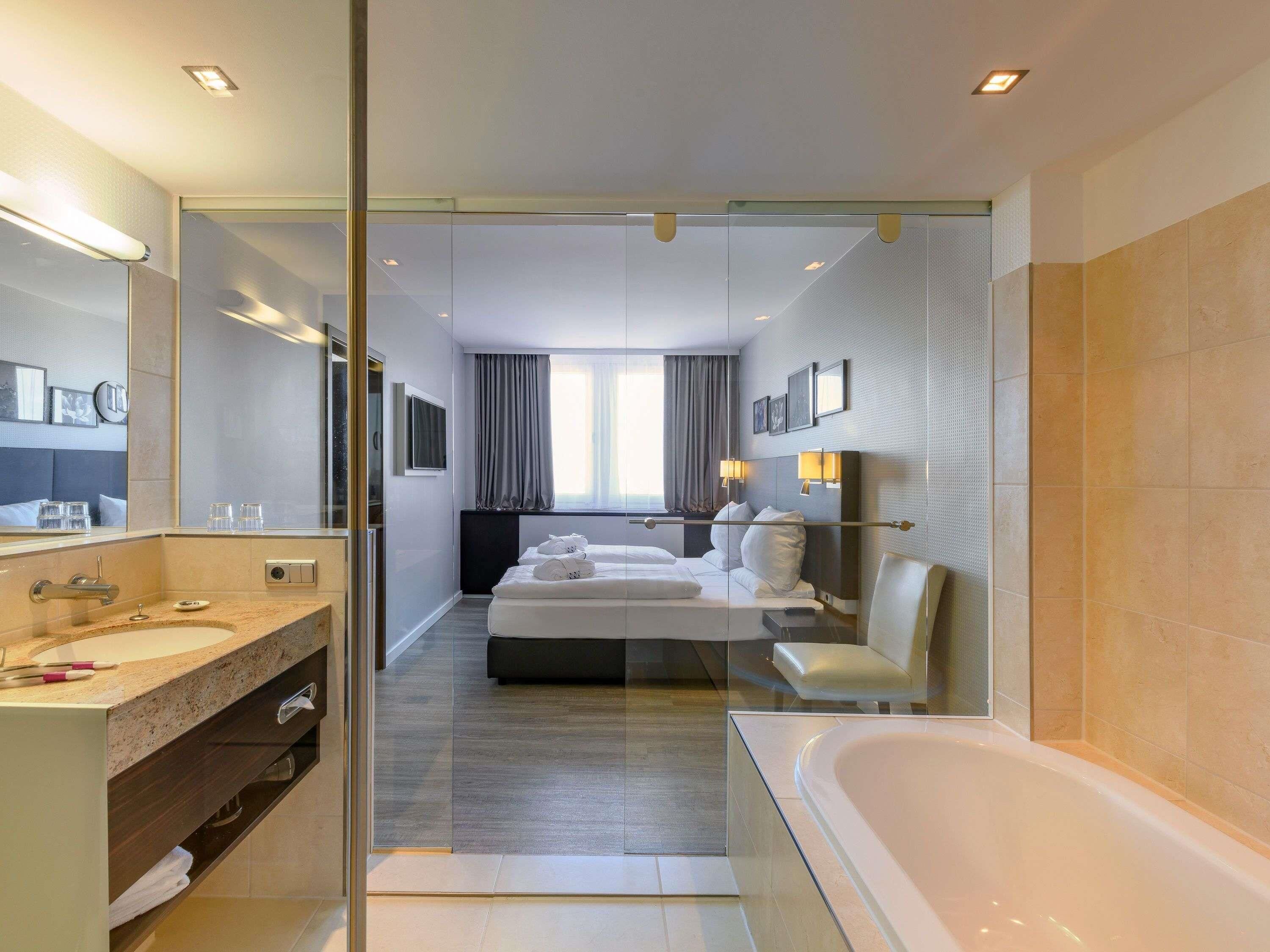Ein modernes Hotelzimmer mit Glastrennwand, einem großen Bett, einem separaten Badezimmer mit Badewanne und einem großen Spiegel.