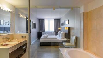 Ein modernes Hotelzimmer mit Glastrennwand, einem großen Bett, einem separaten Badezimmer mit Badewanne und einem großen Spiegel.