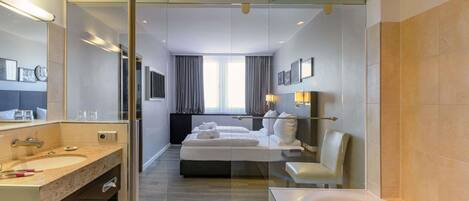 A modern hotel room with a glass partition, a large bed, a separate bathroom with a bathtub, and a large mirror.