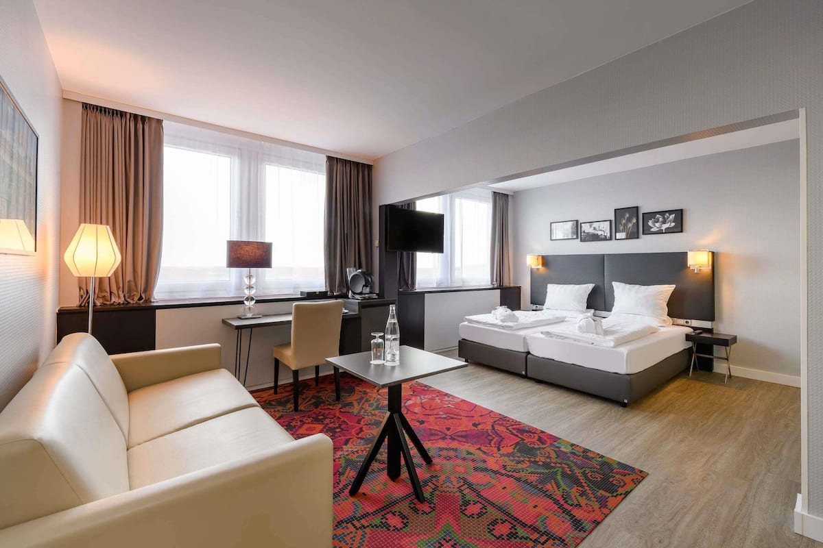 Une chambre d’hôtel comprenant un lit, un bureau, une chaise, un canapé, une télévision et une fenêtre avec des rideaux.