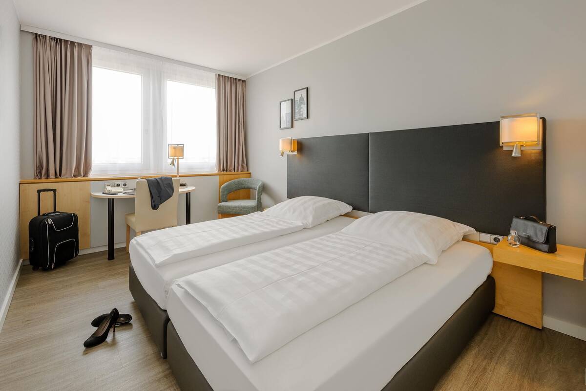 A hotel room with two beds, a desk, a chair, a suitcase, and a lamp.