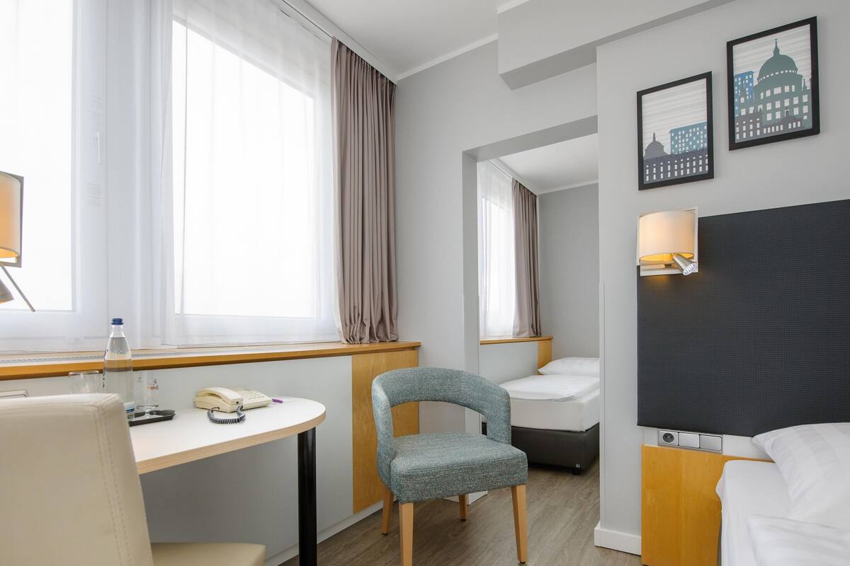 A hotel room with a bed, a desk with a chair, a small table, a lamp, and a window with curtains.