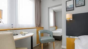 A hotel room with a bed, a desk with a chair, a small table, a lamp, and a window with curtains.