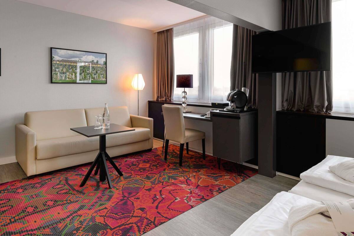 A hotel room with a sofa, a desk, a chair, a bed, a window with curtains, a lamp, and a painting on the wall.