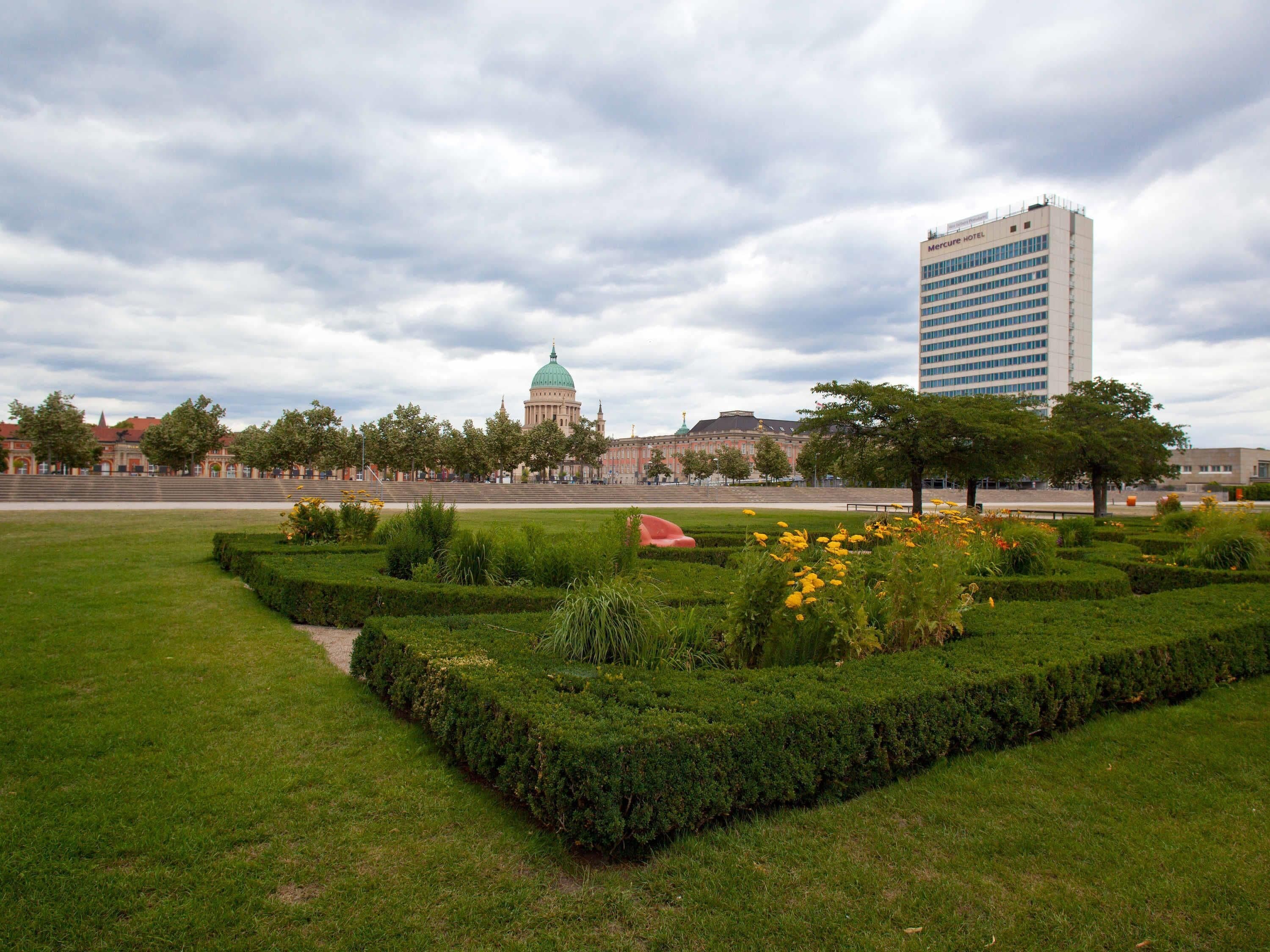 Ein gepflegter Garten mit geschnittenen Hecken und verschiedenen Blumen. Im Hintergrund ein großes Gebäude mit einer Kuppel und einem modernen Hotel.
