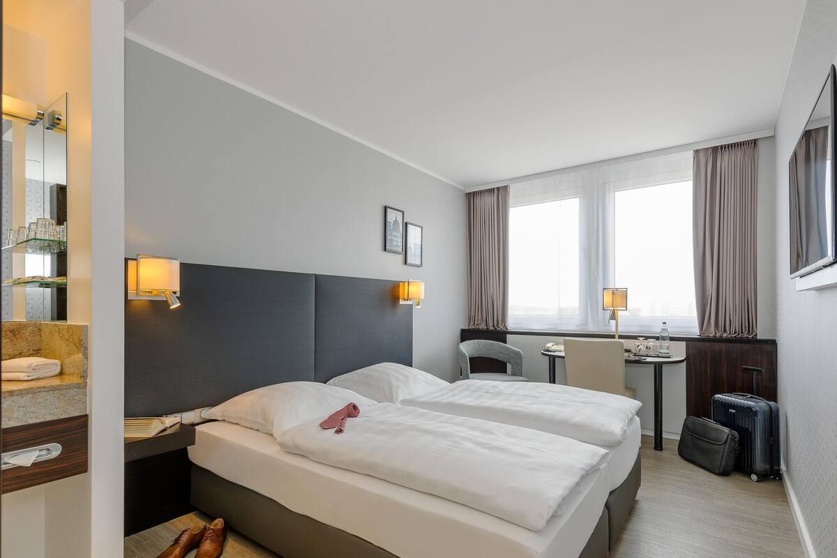 A hotel room with a bed, bedside tables, a desk, a chair, a mirror, and a window with curtains.