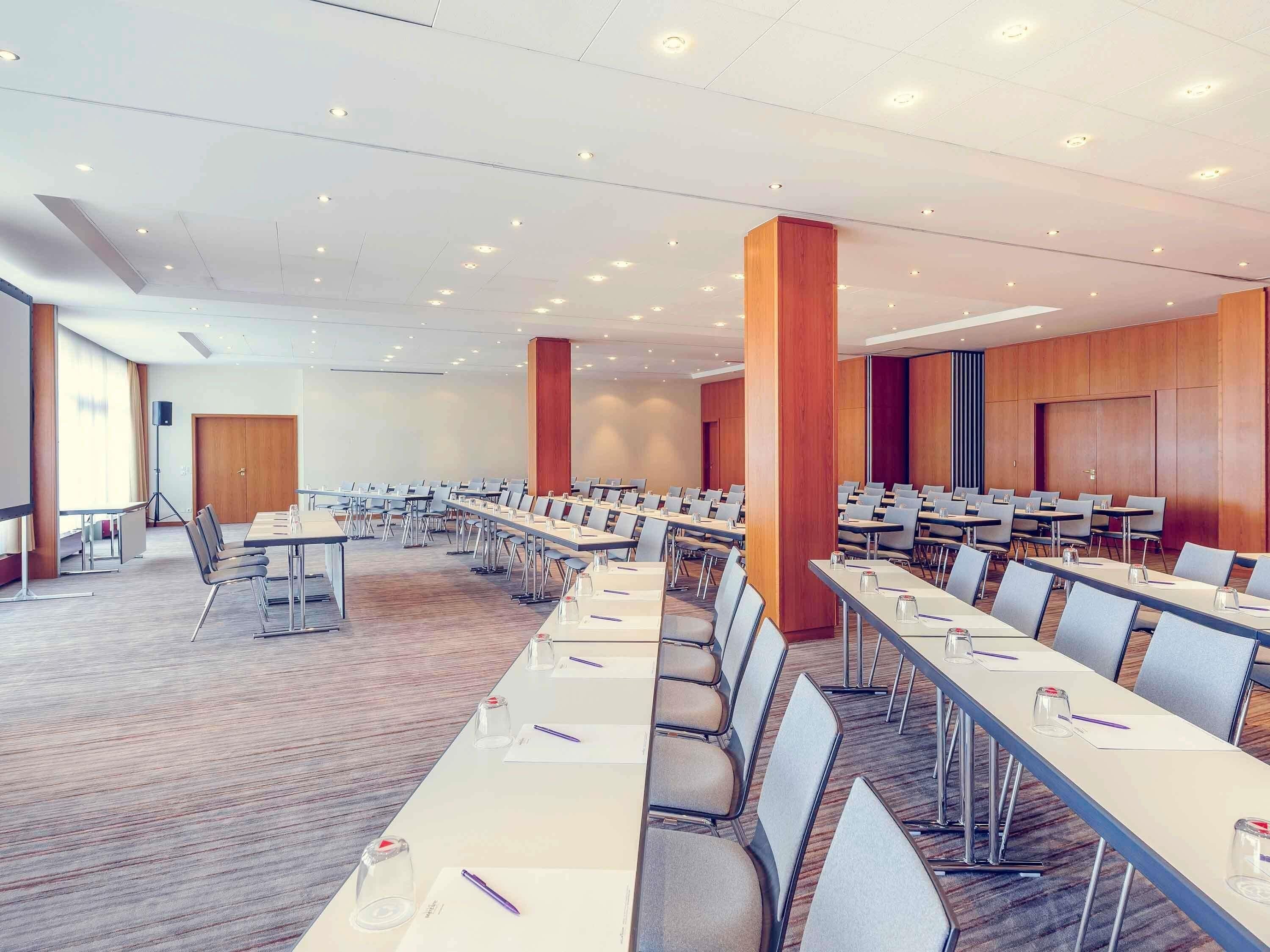 Un salón de conferencias con varias mesas y sillas, paneles de madera e iluminación empotrada en el techo.