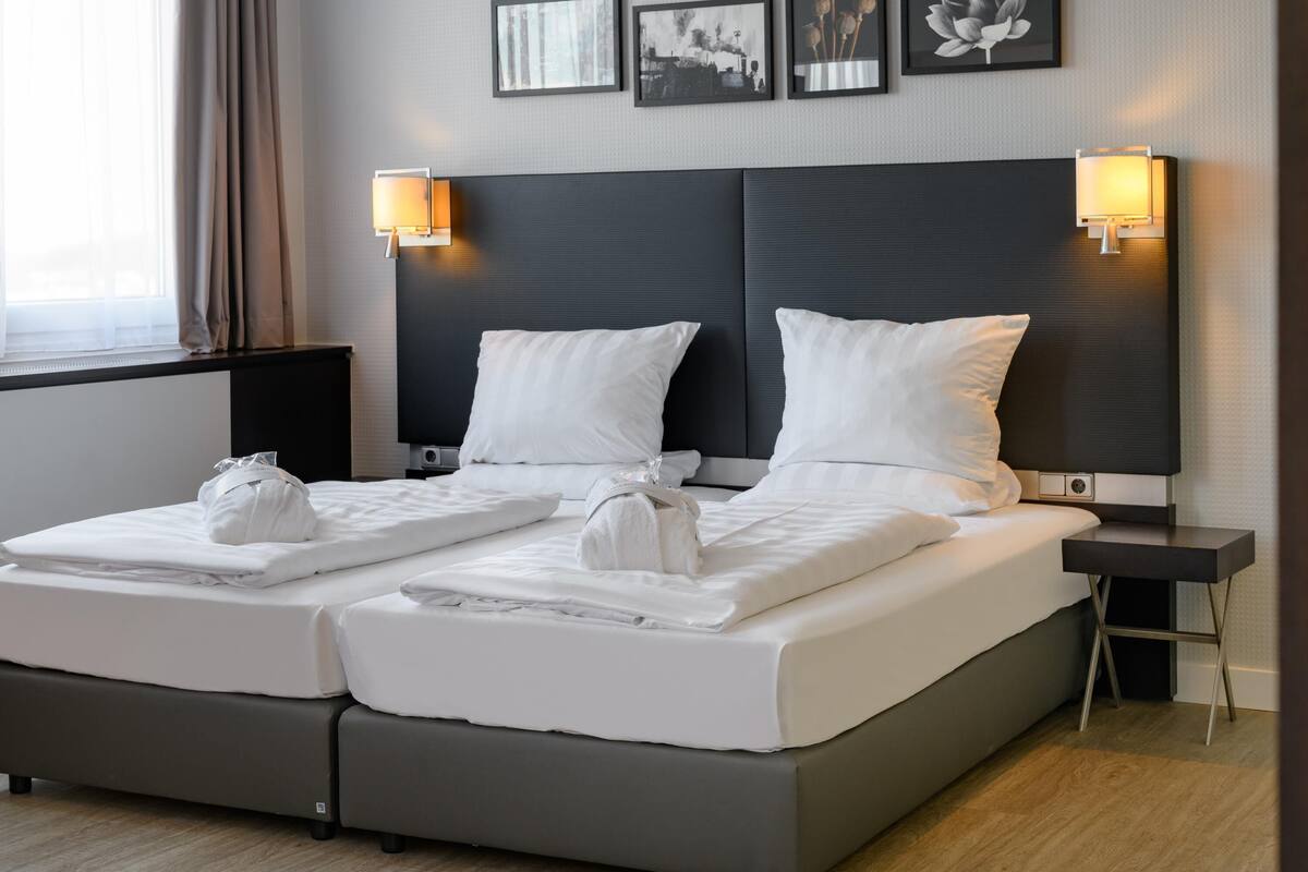 A double bed with two pillows, a bedside table, a wall-mounted lamp, and a window with curtains.