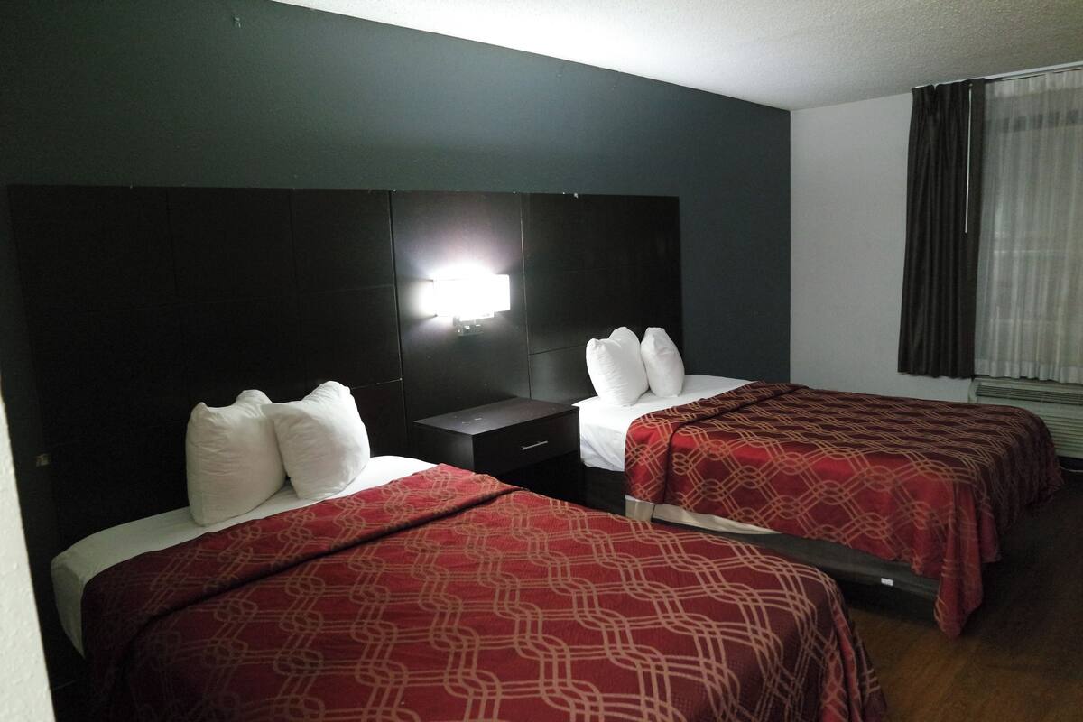 A hotel room with two beds, each with a red patterned bedspread and white pillows. There is a nightstand between the beds, and a wall-mounted light above the left bed.