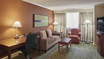 Habitación de hotel con pared de madera, un sofá, un escritorio con lámpara, una silla, un televisor y suelo alfombrado.