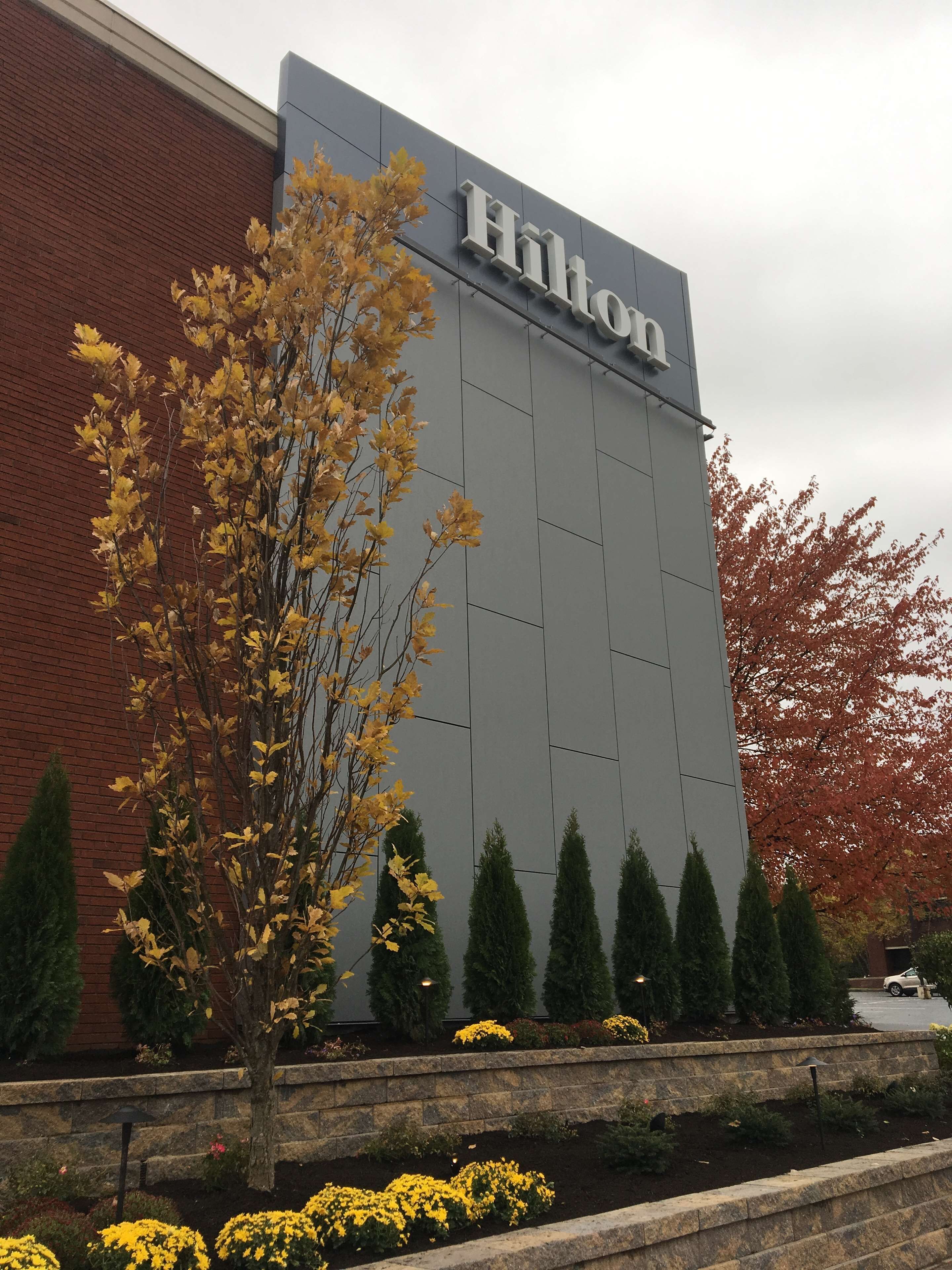 Een Hilton hotelbordje met een bakstenen gevel en een aangelegde tuin met gele bloemen en bomen.